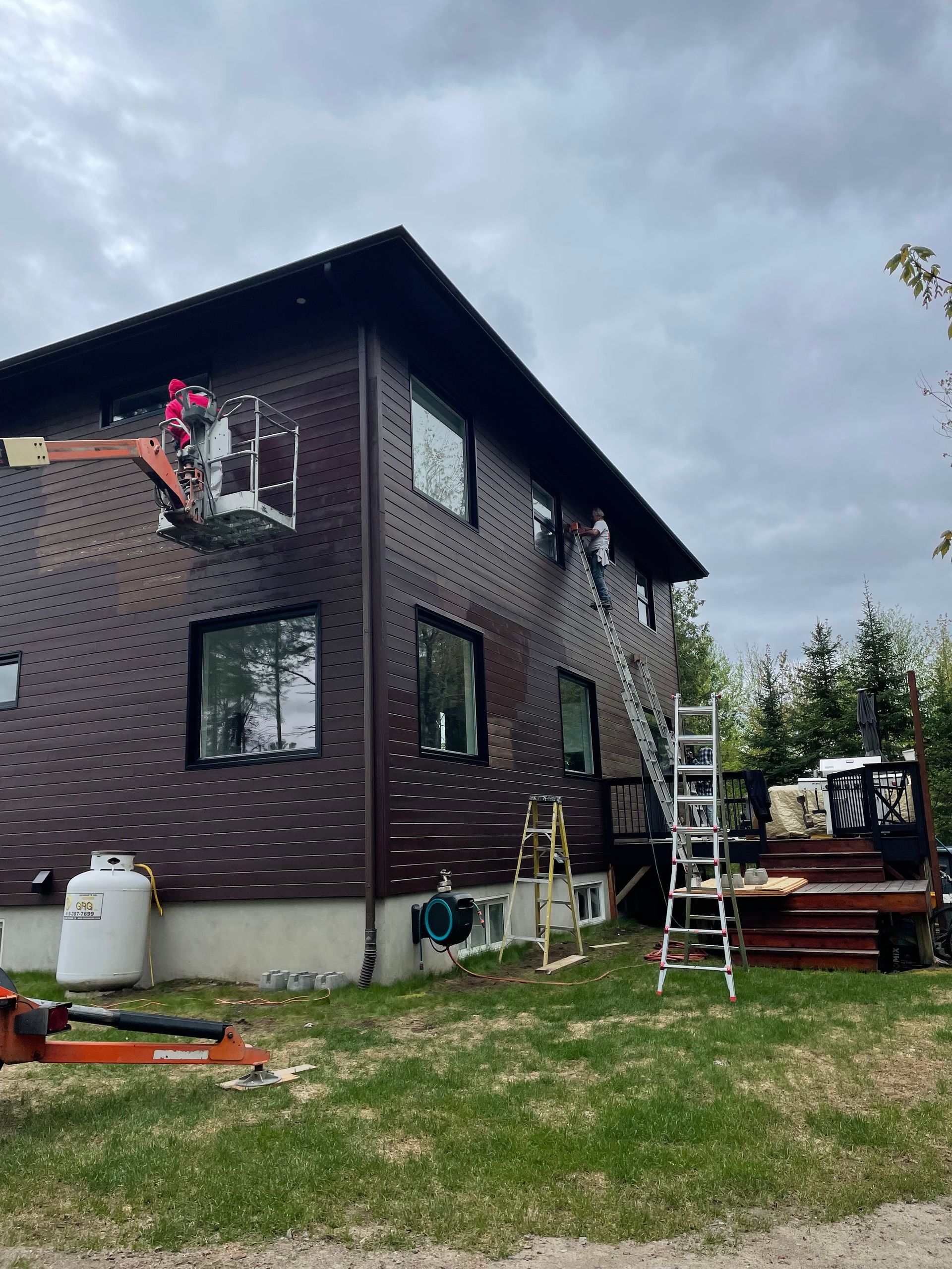 Des ouvriers, perchés sur des nacelles et des échelles, installent des fenêtres sur un bâtiment brun de deux étages avec une terrasse, par temps nuageux.