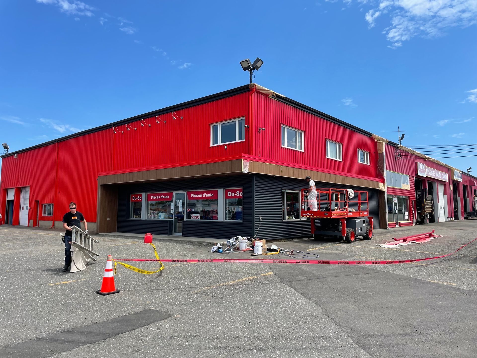 Bâtiment commercial rouge et noir avec une personne debout près d'une échelle et d'un ascenseur ; travaux en cours.
