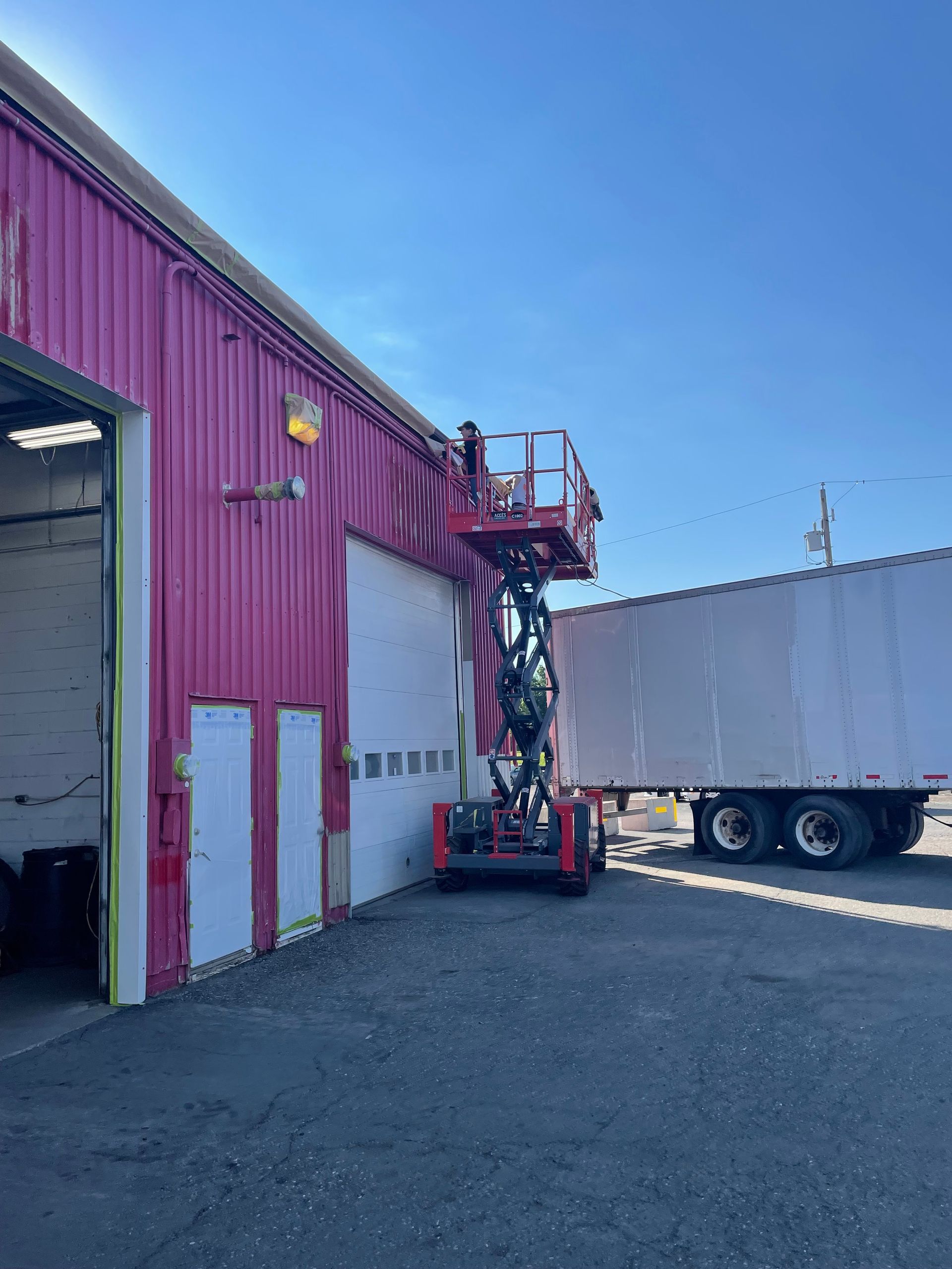 Une personne se trouve sur une nacelle élévatrice rouge près d'un bâtiment rose en train de peindre sa façade. Un semi-remorque est stationné à proximité.