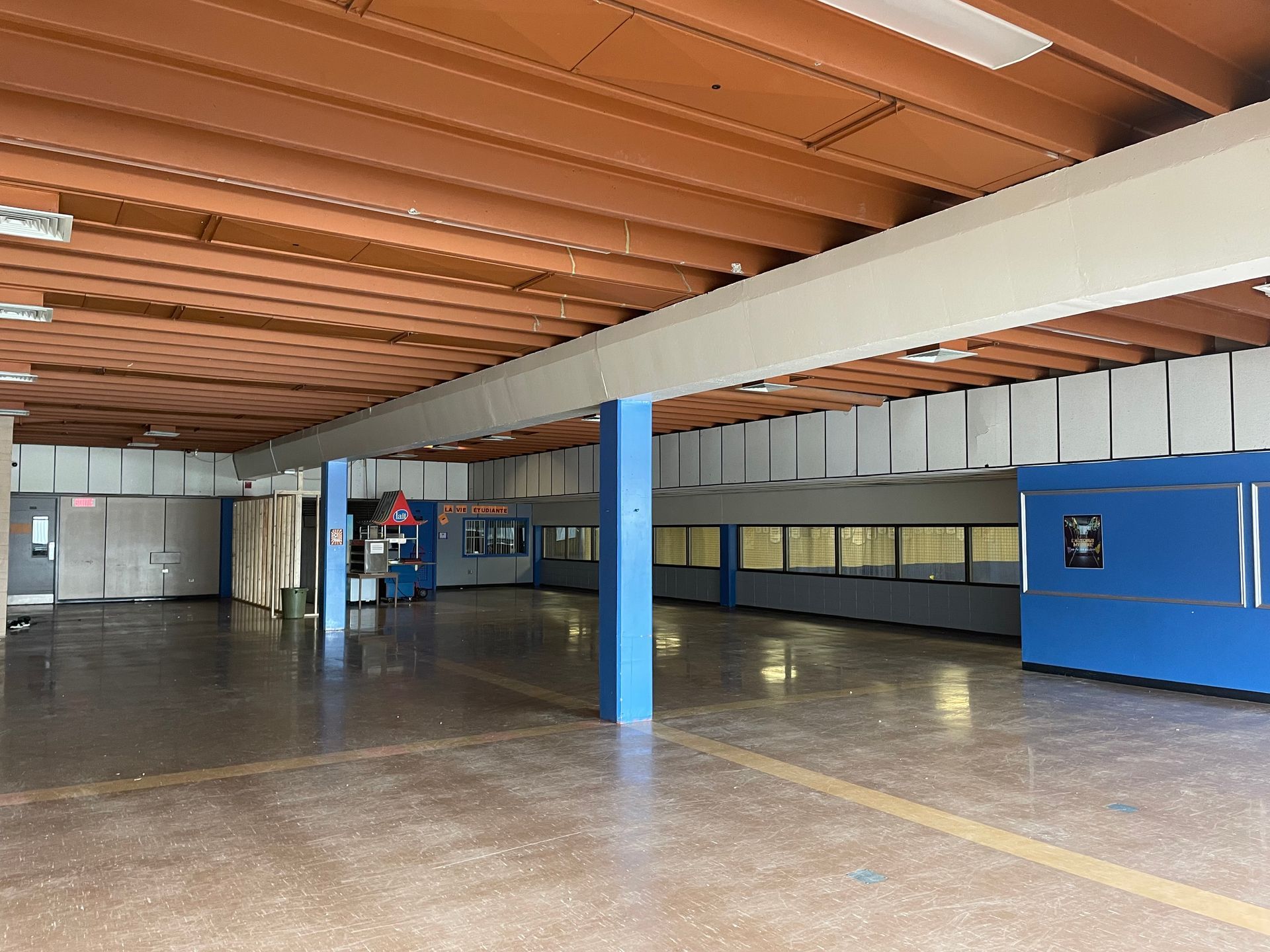 Espace intérieur vide et ouvert avec des piliers bleus, des poutres de plafond brunes et un sol en béton.
