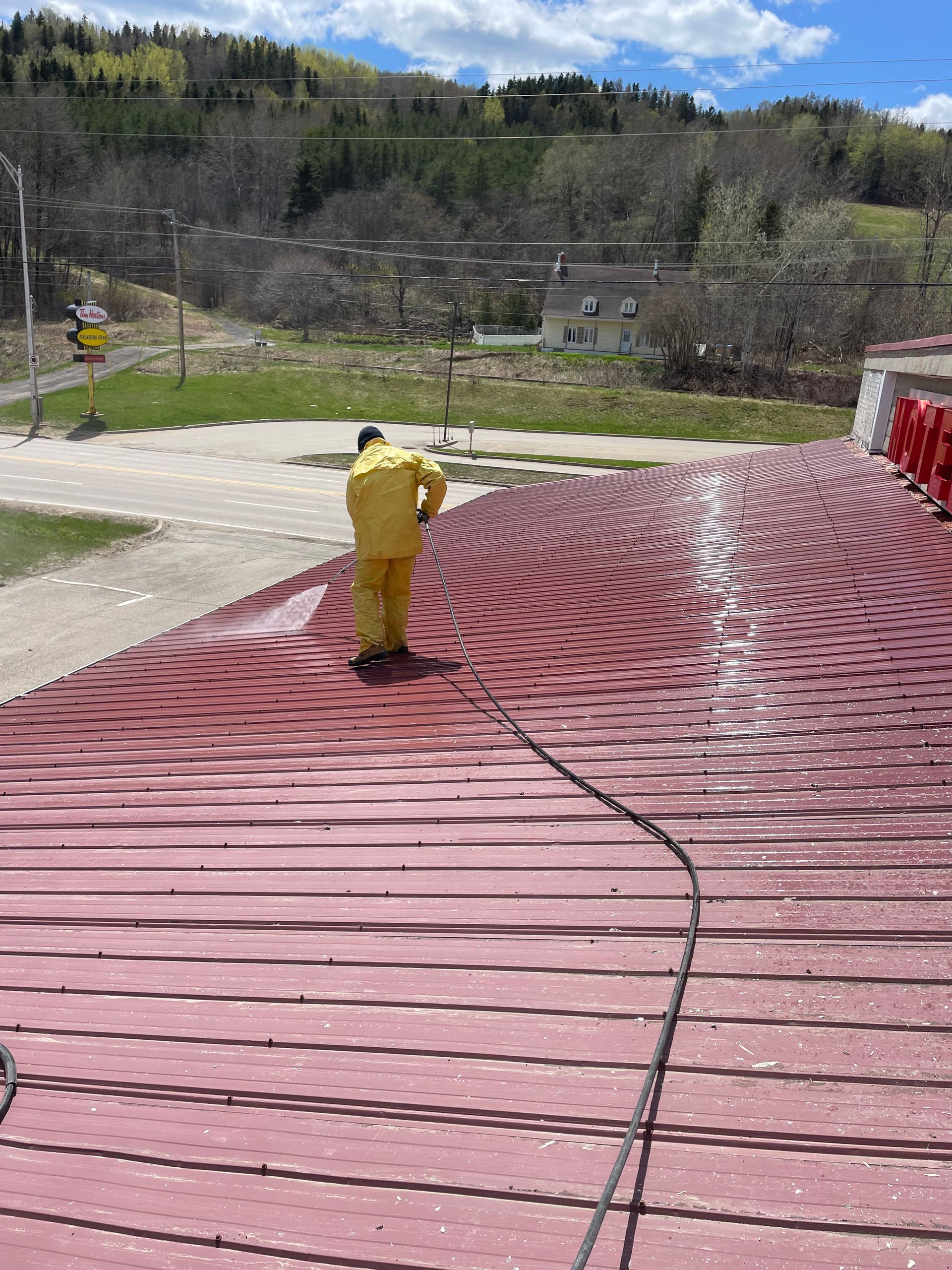 Personne en combinaison de protection jaune nettoyant au jet d'eau un toit en métal rouge à l'extérieur.