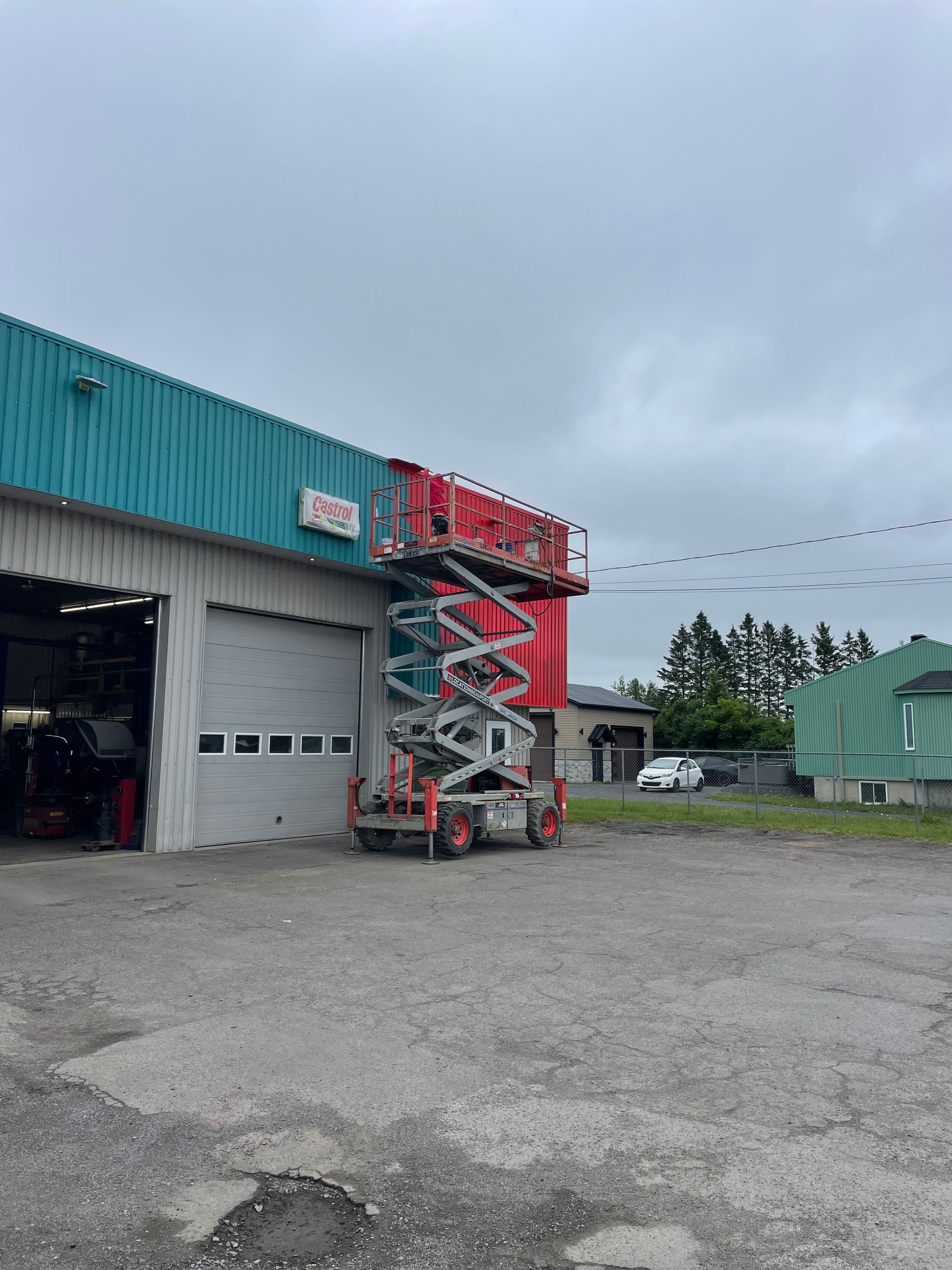 Nacelle élévatrice rouge devant un bâtiment turquoise avec une porte de garage, par temps nuageux.