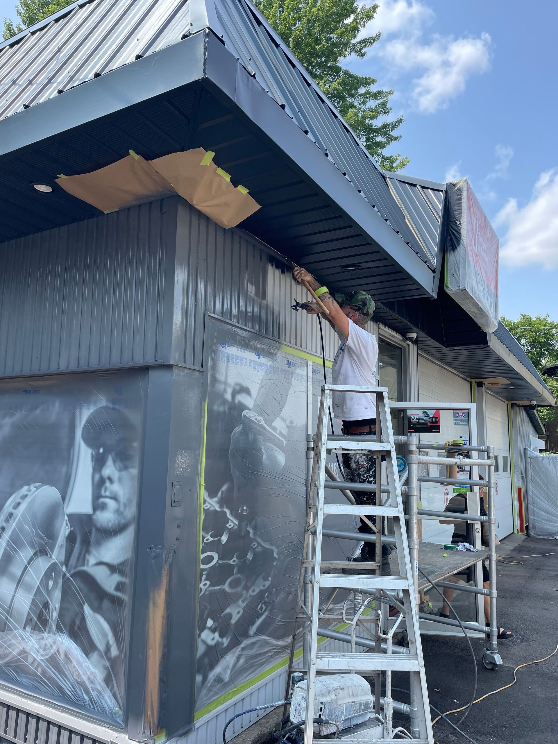 Une personne peint une fresque sur la façade d'un bâtiment. Elle se tient sur une échelle, des échafaudages se trouvant à proximité.