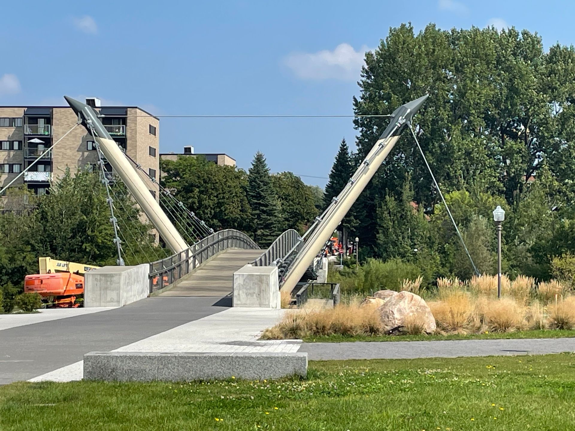 Une passerelle piétonne aux hauts piliers inclinés. Un chemin en béton mène à un pont arqué enjambant un espace vert. Des bâtiments se dressent à l'arrière-plan.