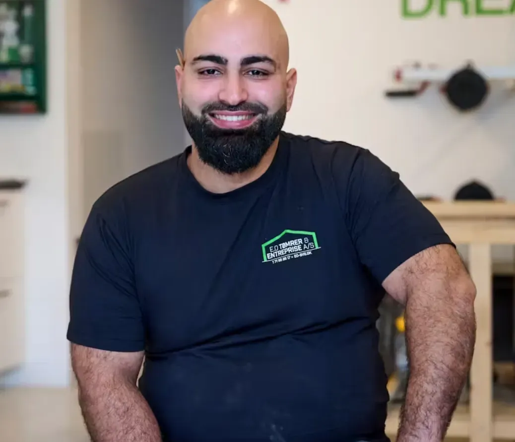 Skaldet mand med fuldskæg smiler, iført en sort t-shirt med logo. Indendørs, sandsynligvis i en forretning.