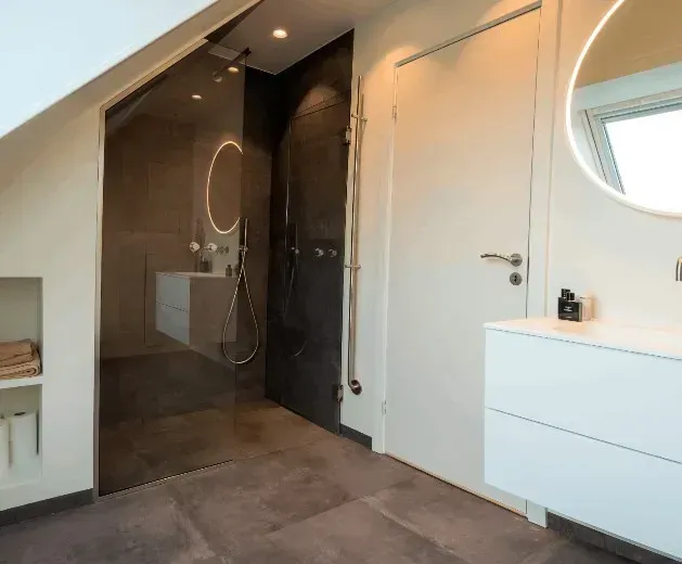 Moderne badeværelse med glasbruser, hvid håndvask og rundt spejl.