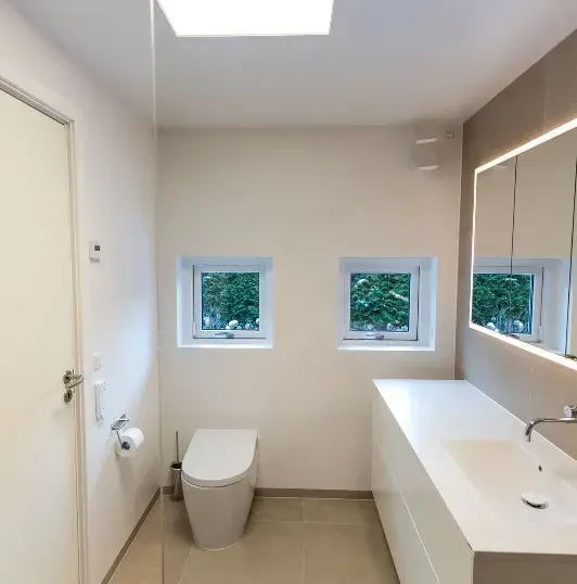 Moderne badeværelse med hvide vægge, to små vinduer og et toilet.