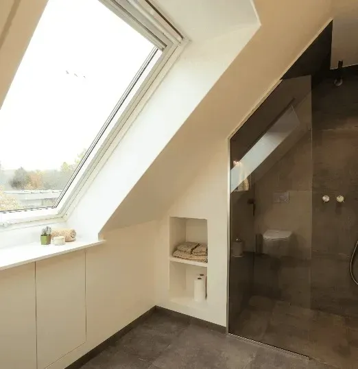 Badeværelse med vinklet væg, ovenlysvindue, bruser, indbygget opbevaring og gråt flisegulv.