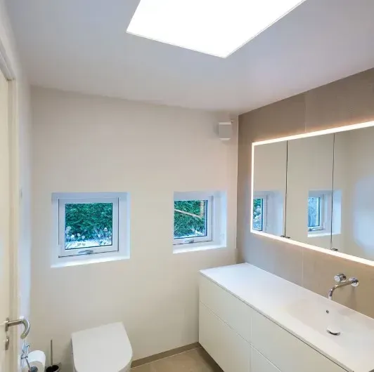 Moderne badeværelse med ovenlysvindue, to små vinduer, hvidt håndvask og et baggrundsbelyst spejl.