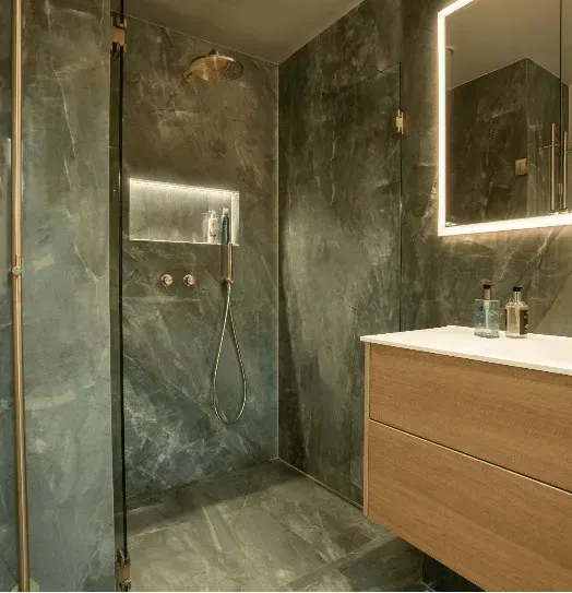 Moderne badeværelse med grå marmorvægge, håndvask i træ og baggrundsbelyst spejl. Bruseområde med messingarmaturer.