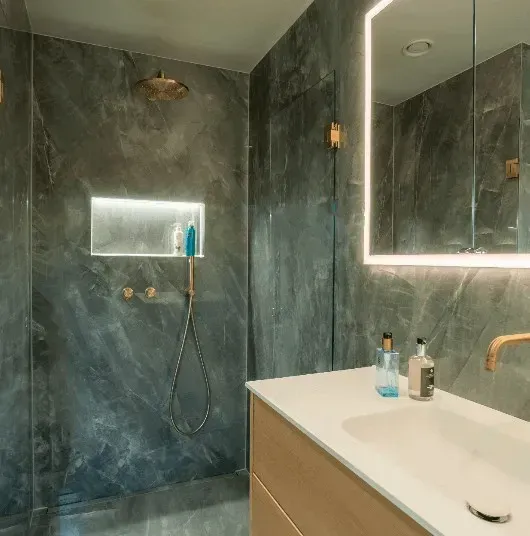 Moderne badeværelse med grå marmorbruser, guldarmaturer og et oplyst spejl.
