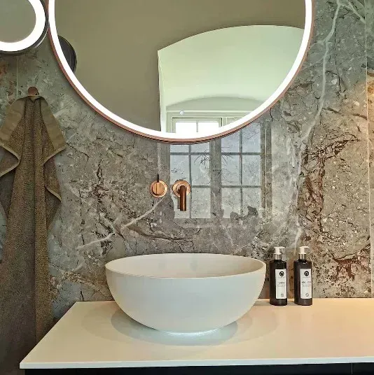 Badeværelse med rundt spejl, håndvask og grå stenlignende væg. Et håndklæde hænger til venstre.