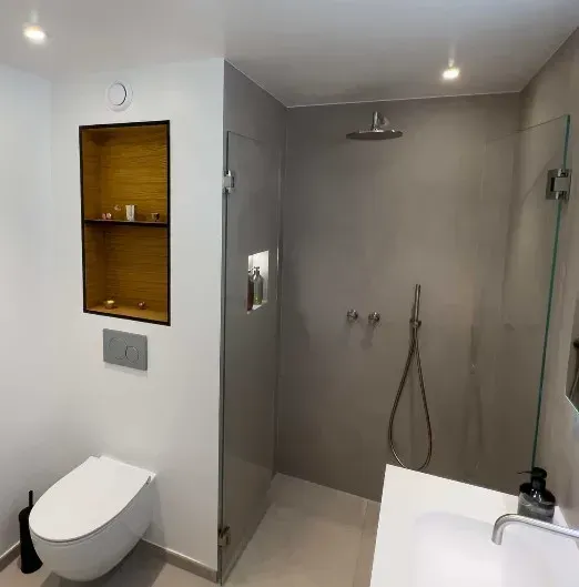 Moderne badeværelse med grå bruser, hvidt toilet og træhylde.