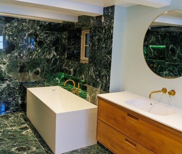 Badeværelse med smaragdgrønne marmorvægge og -gulv, hvidt rektangulært badekar, træbordplade og guldarmaturer.