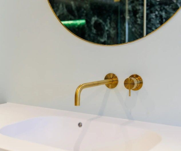Gyldne vandhane og betjeningselementer på hvid væg over en hvid vask med et rundt spejl med guldindramme.