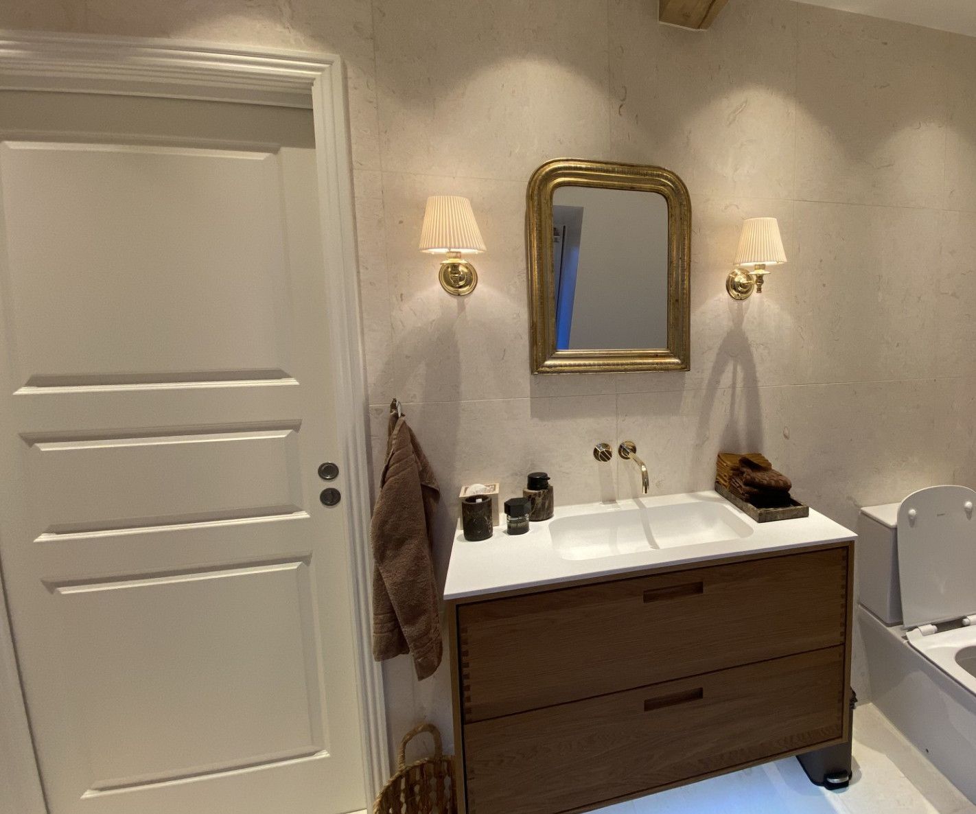 Badeværelse med en hvid dør, spejl med guldindramme, træbordplade og to lampetter.