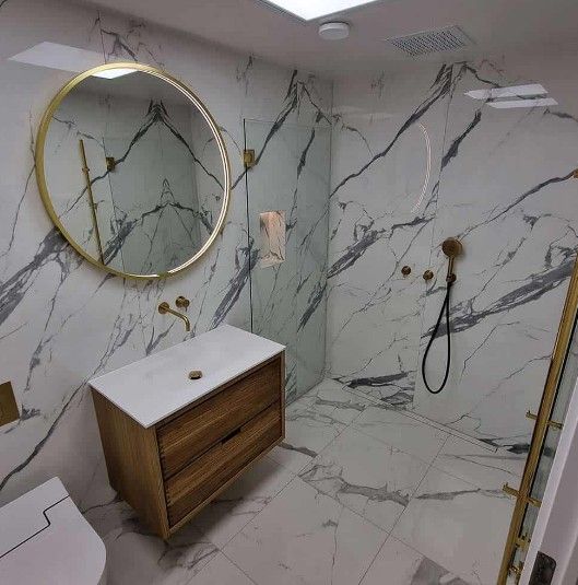 Badeværelse med marmormønstrede vægge og gulv, guldarmaturer, træbordplade og et rundt spejl.