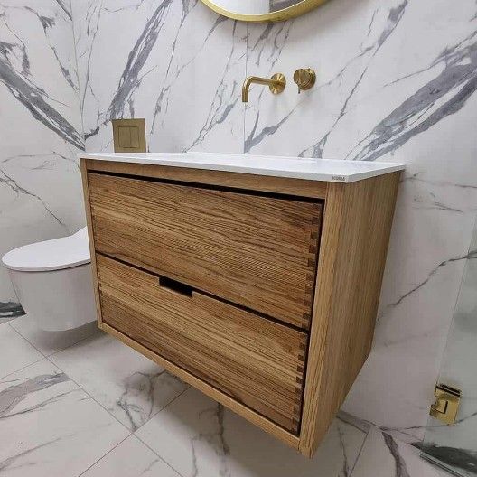 Svævende badeværelsesmøbel i træ med to skuffer, hvid bordplade, guldfarvede armaturer og marmorfliser.