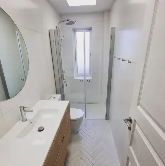 Hvidt badeværelse med bruser, toilet og håndvask. Sildebensflisebelagt gulv.