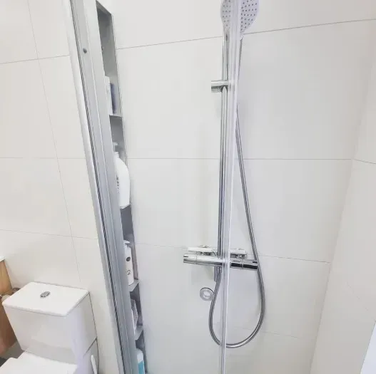 Bruser med forkromede armaturer og skydedør i glas ved siden af et højt, smalt opbevaringsskab.