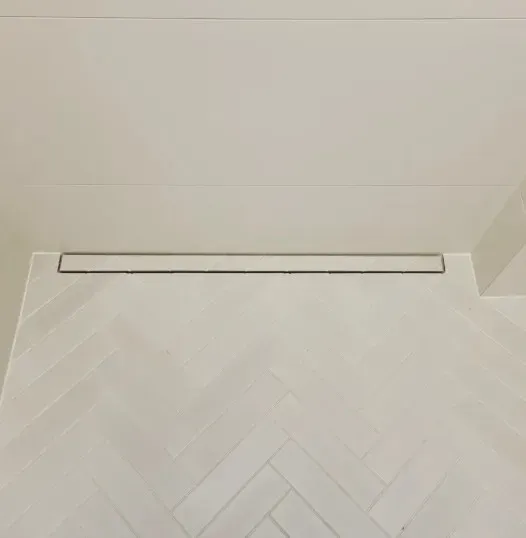 Et langt, rektangulært afløb i et hvidt flisebelagt gulv med et sildebensmønster. Afløbet er placeret tæt på en hvid væg.