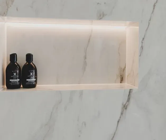 Indbygget bruseniche med to mørke flasker. Blød belysning oplyser marmorfliser.