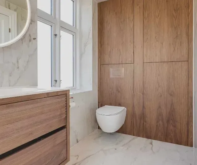 Badeværelse med træpaneler, vægmonteret toilet og marmorbadeværelse med vindue.