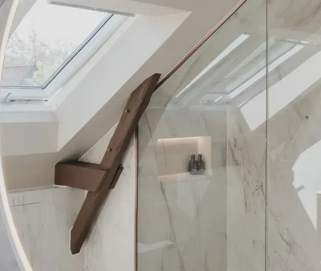 Badeværelse med glasbrusekabine, marmorvægge, ovenlysvindue og træbjælkedetaljer.