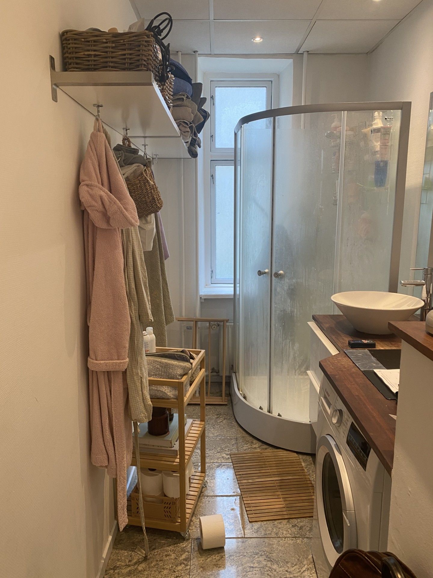 Lille badeværelse med bruser, vaskemaskine, hylder med håndklæder og vindue.