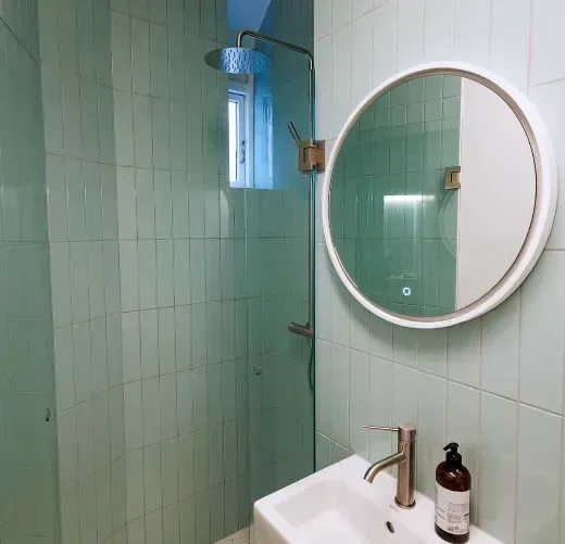 Grønt flisebelagt badeværelse med bruser, rundt spejl og håndvask.