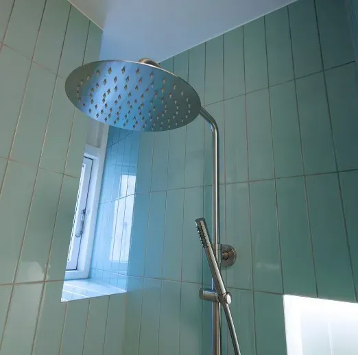 Sølvbruserhoved og håndbruser mod blågrøn flisebelagt væg, nær et vindue.