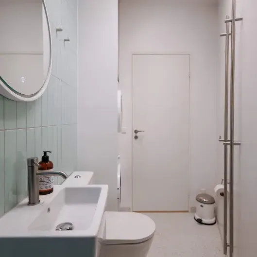 Badeværelse med hvid vask, toilet og dør. Pastelblå fliser på væggen og et spejl.