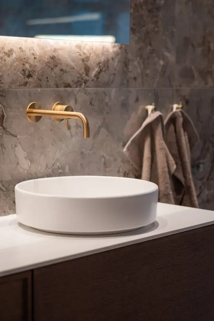Hvid vask og gylden vandhane på et badeværelsesbord med håndklæder; placeret op ad en marmorvæg i et badeværelse.