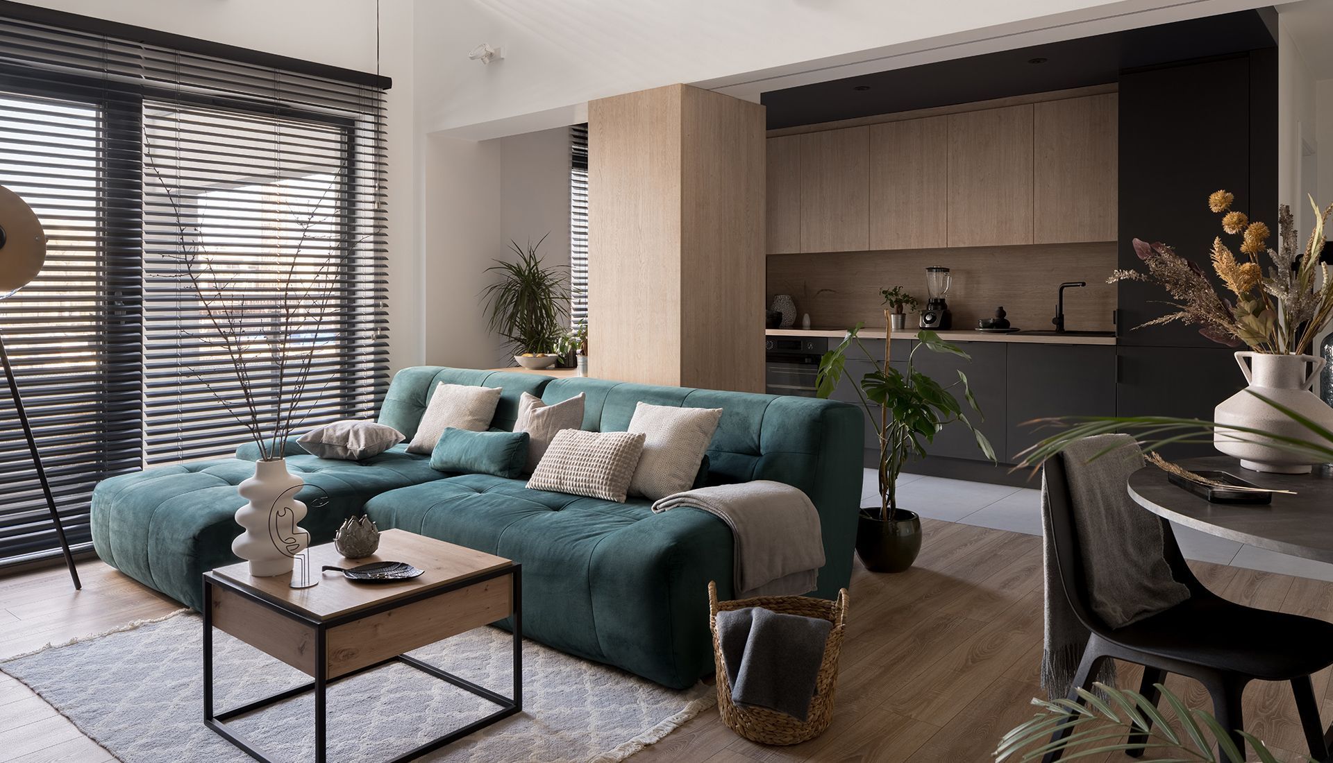 Teal sectional sofa in living room with a kitchen in the background. Wooden floors, black blinds, neutral tones.