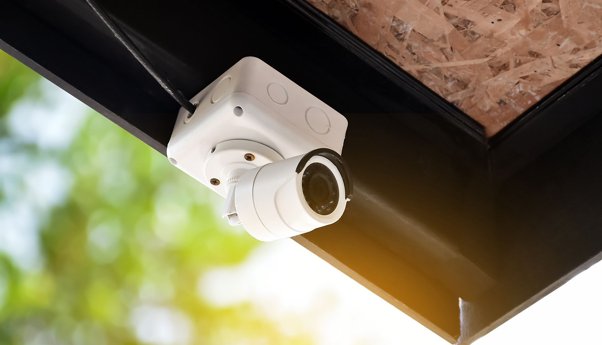 White security camera mounted on a black overhang, with a blurred green and gold background.