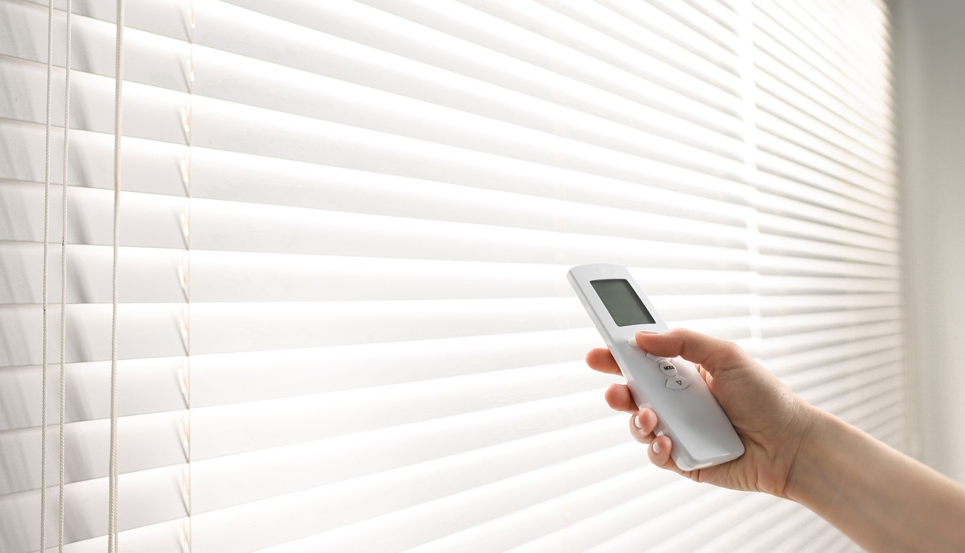 Hand holding a remote control adjusting white, horizontal blinds.