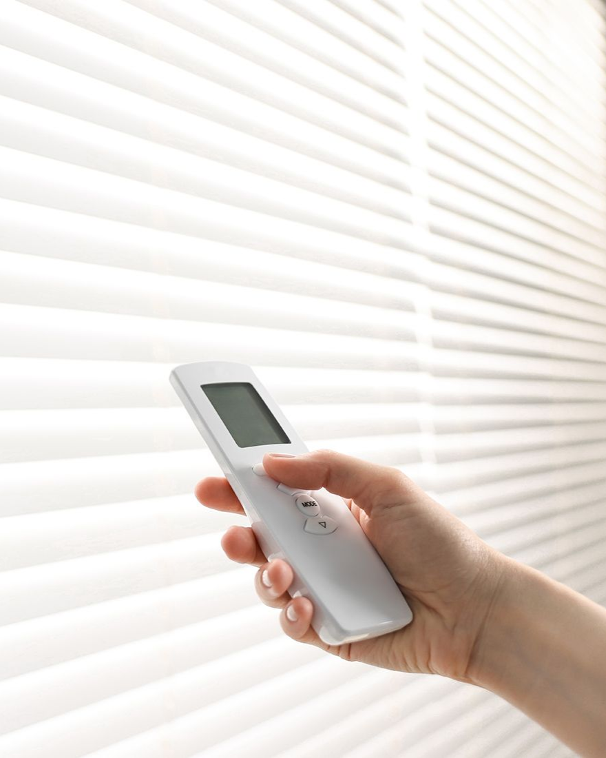 Hand holding a white remote control, adjusting white window blinds.