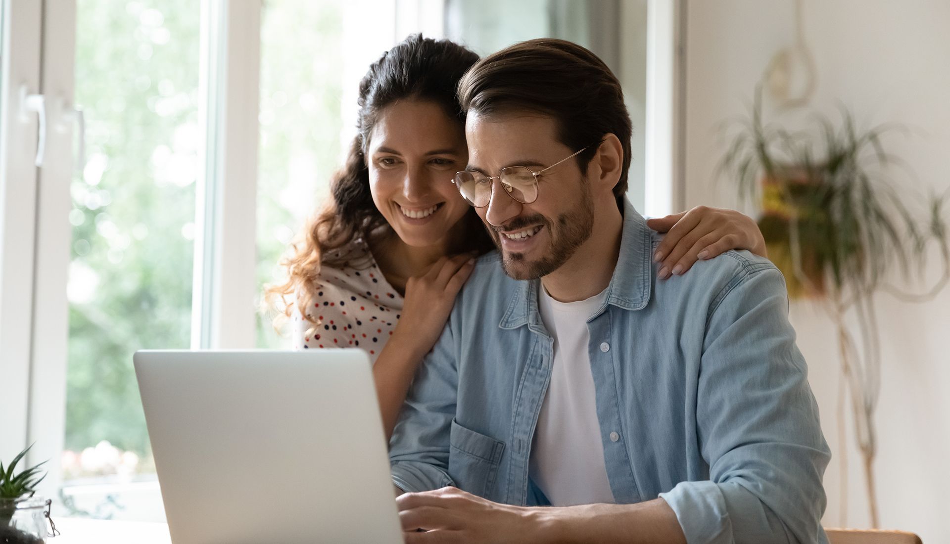 Couple smiling at a laptop, woman with arm around man. Bright room with window.