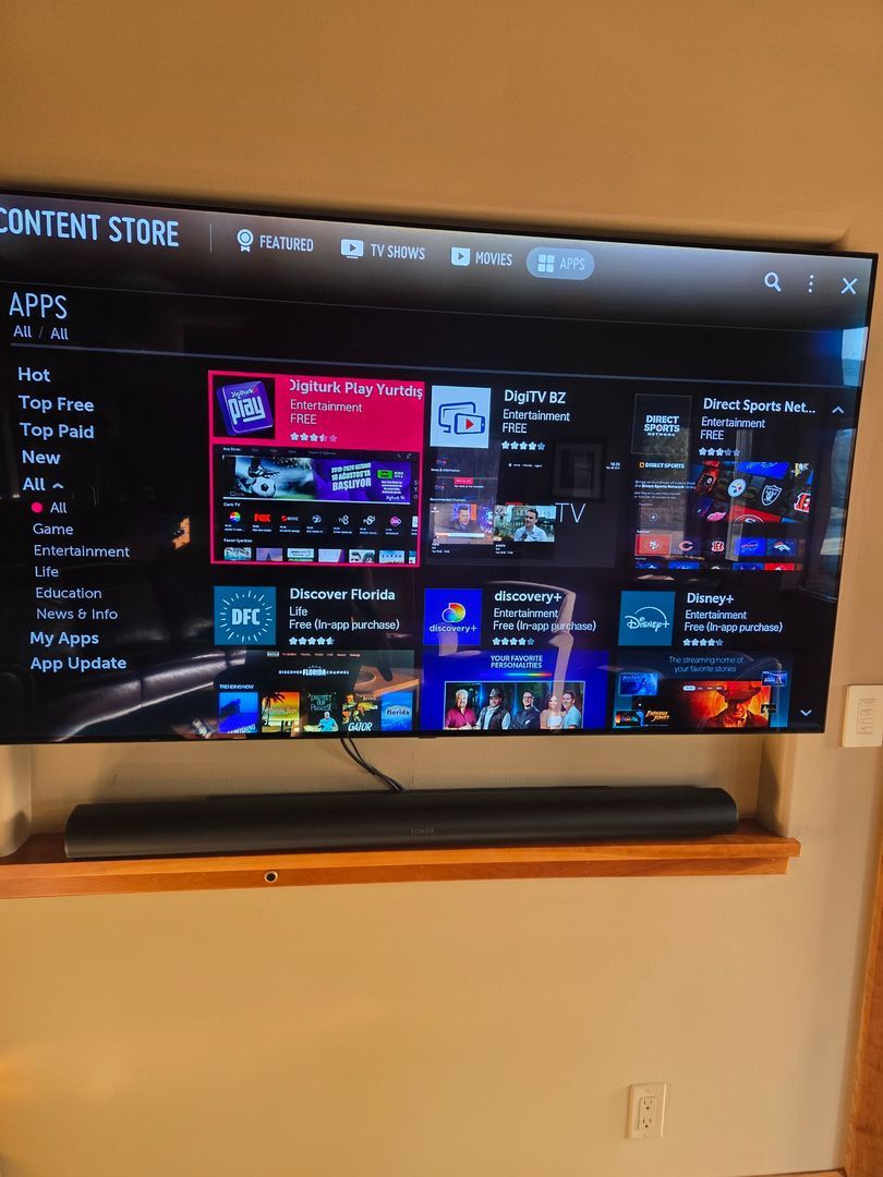 A wall-mounted TV displays an app store interface with a black soundbar on a wooden shelf beneath it.
