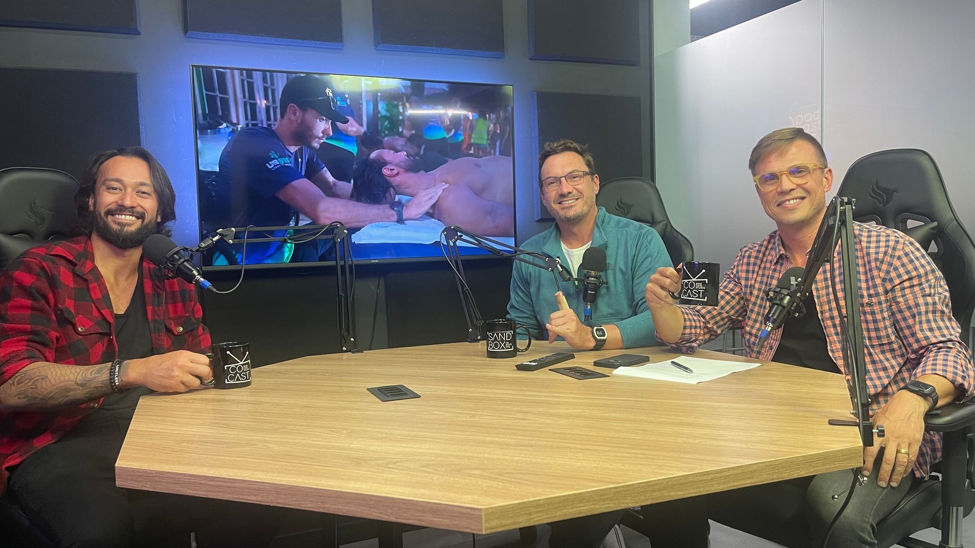 Three men at a table in a studio, smiling into microphones. TV screen in the background.