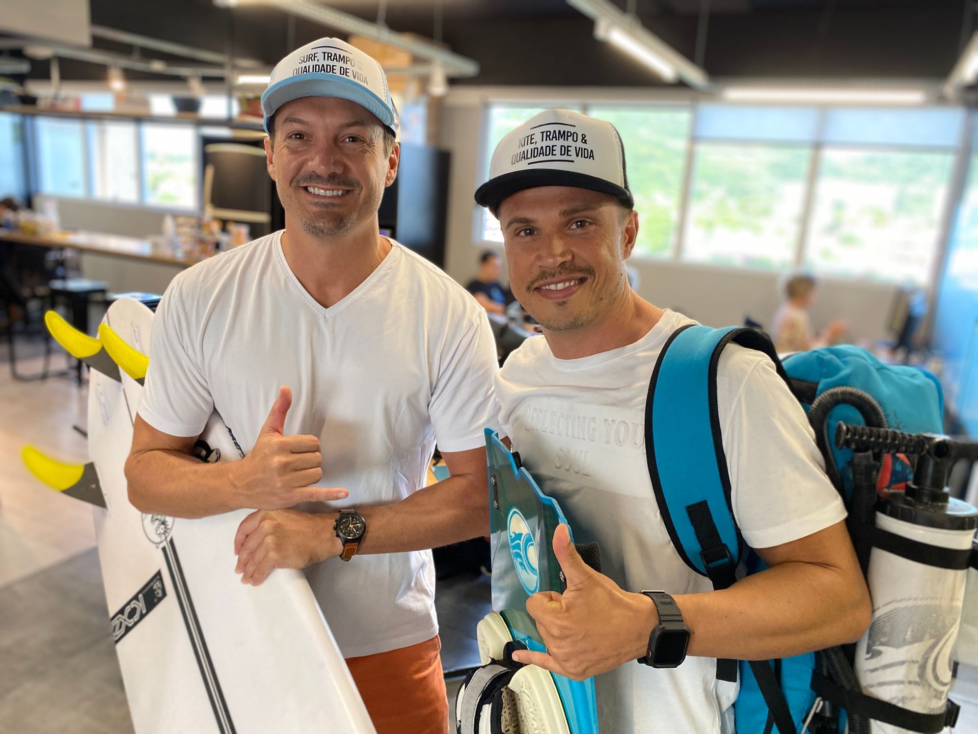 Dois homens segurando pranchas de surfe, sorrindo e fazendo sinal de positivo. Em ambiente interno, usando chapéus, um deles com mochila.