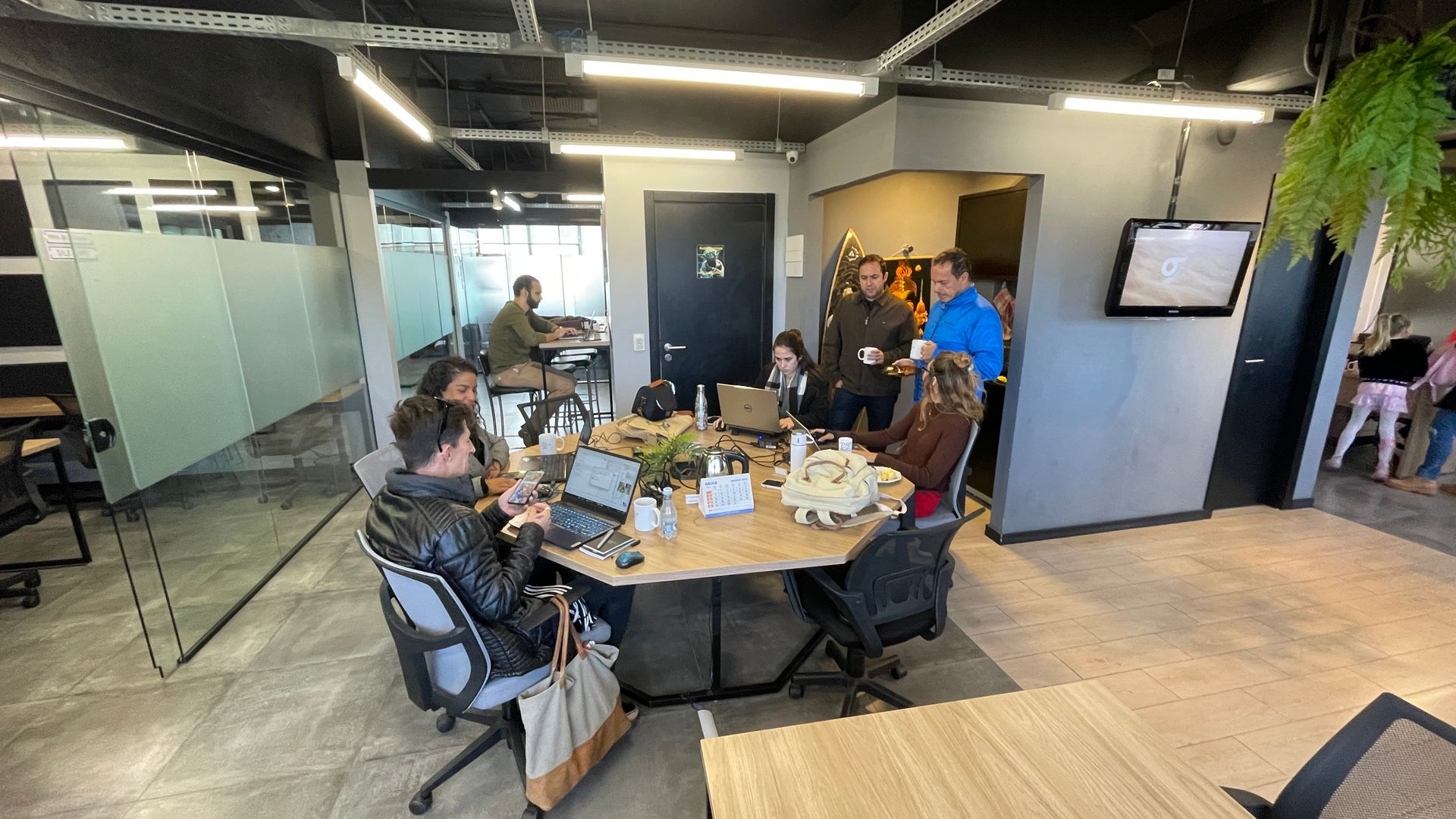 A group of people in an office setting around a table, using laptops. Some are standing and talking.