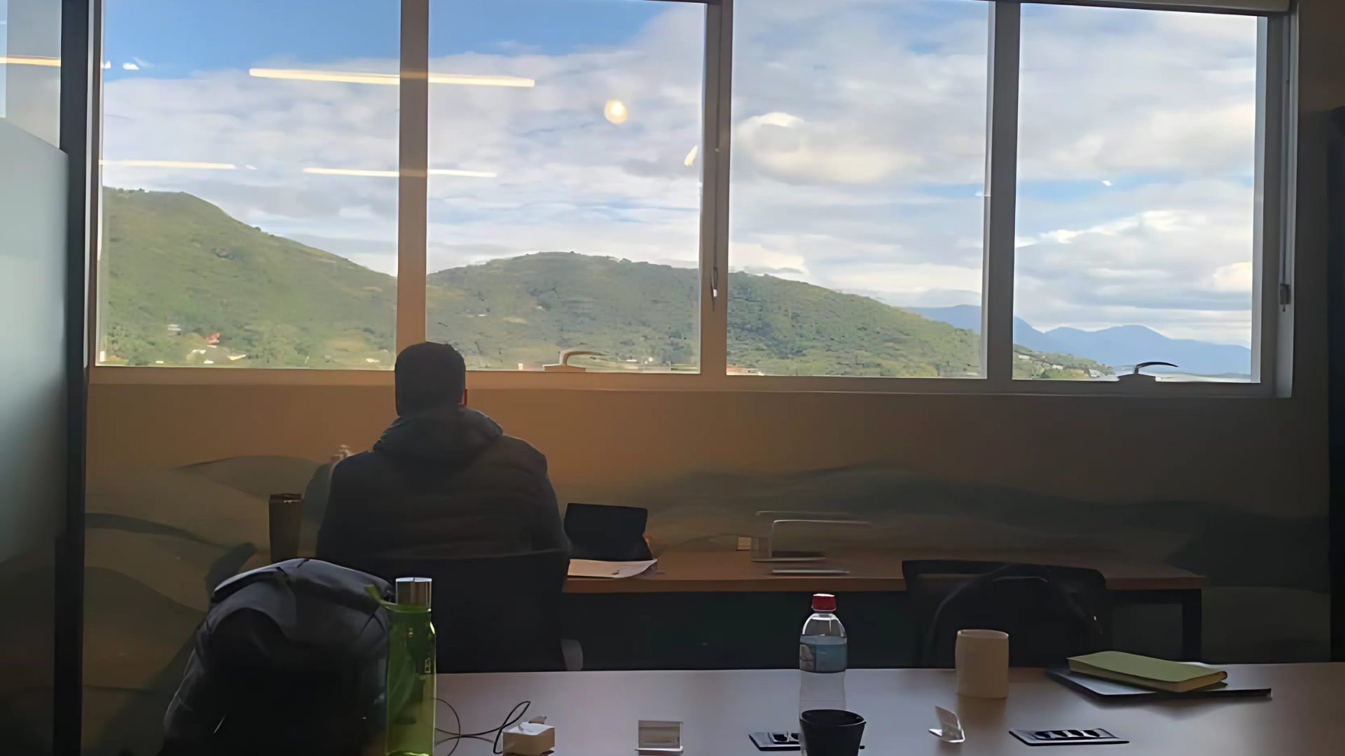 Um homem está sentado à mesa em frente a uma janela com montanhas ao fundo.