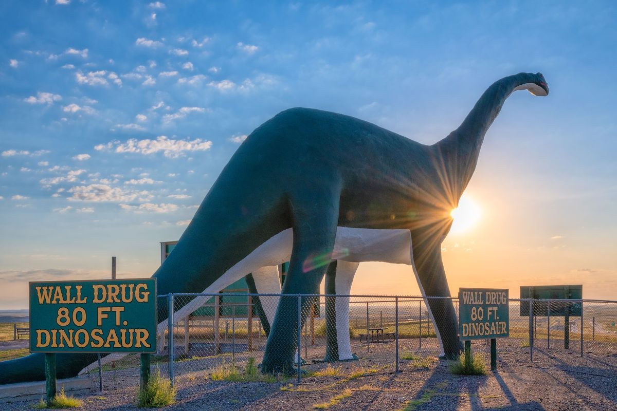 A giant dinosaur is standing next to a sign that says wall drug 80 ft dinosaur.