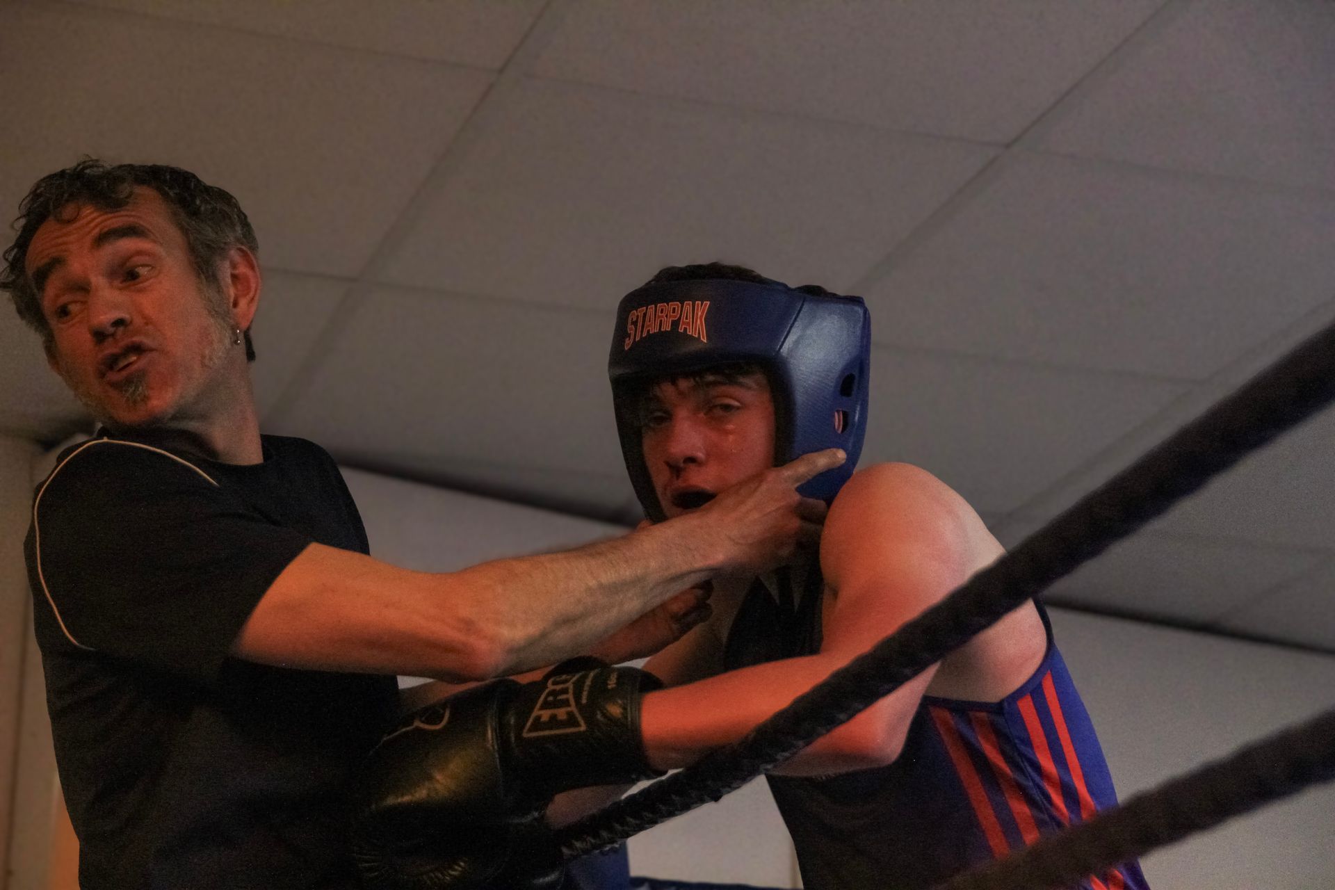 Boxer in ring, coach adjusting headgear, looking concerned. Black gloves, blue and red attire, setting is boxing gym.