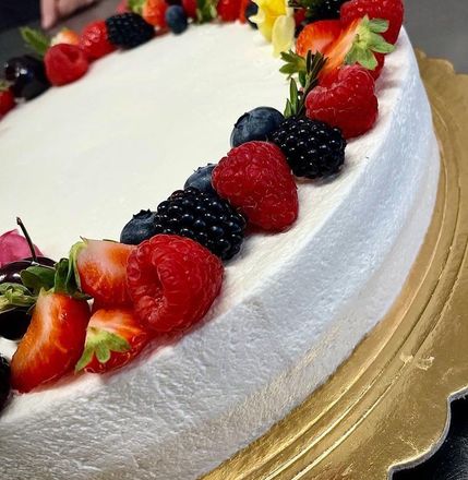 Torta glassata bianca decorata con fragole, lamponi, mirtilli e more su un vassoio dorato.