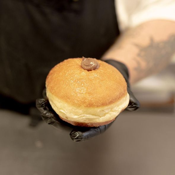 Una persona che indossa guanti neri tiene in mano una ciambella dorata con un ciuffo di ripieno di cioccolato sopra.