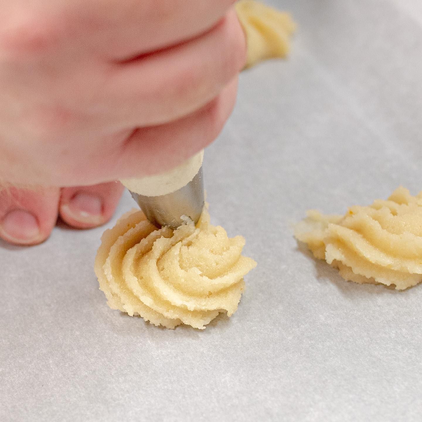 Una persona che con una sac à poche versa l'impasto dei biscotti su carta da forno, creando delle forme a spirale.