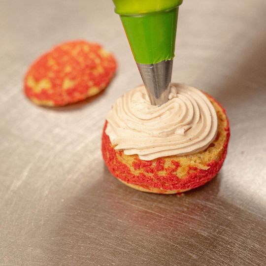 A person is spreading hazelnut-coloured cream on a cake with a red surface. Another cake can be seen in the background.