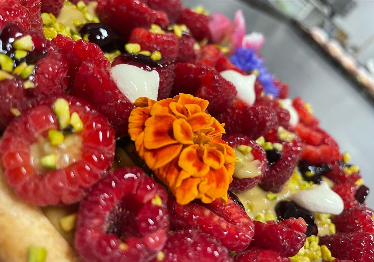 Primo piano di una crostata dolce guarnita con lamponi freschi, pistacchi, panna e un vivace fiore arancione.